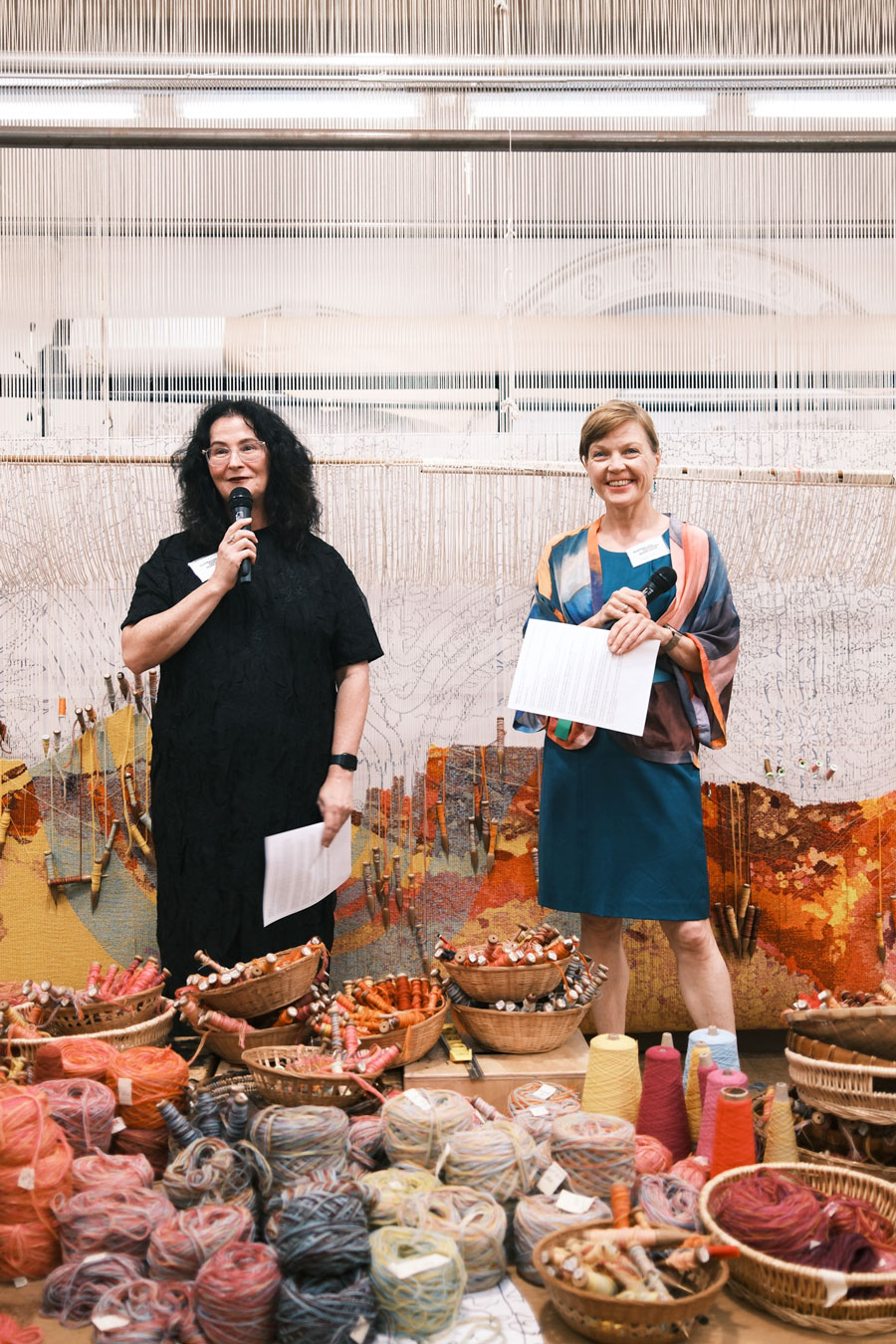Two women stand in front of a loom, piles of colourful yarn in front of them