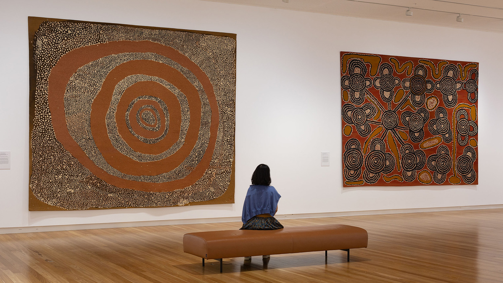 A woman sits in the gallery space at Bunjil Place looking at two tapestries included in the 'Woven Together' exhibition. 
