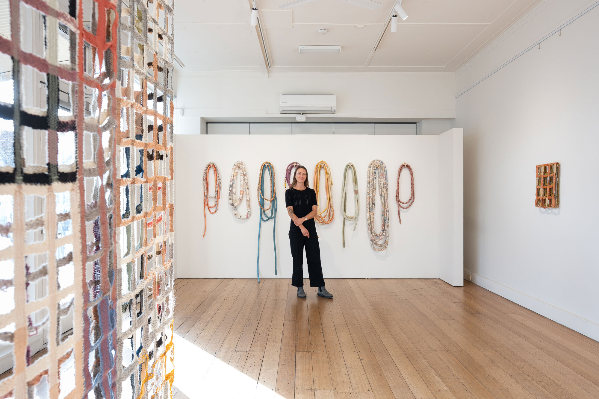‘Entanglement’, 2025, Kayla Powers. Photo: Astrid Mulder A woman stands in a gallery space amongst her textile artworks, some artworks resemble nets or trellises, while others look like long sausages looped around. The colours are muted.