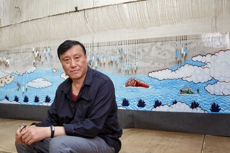 Guan Wei in front of 'Treasure Hunt' 2018, designed by Guan Wei and woven by Chris Cochius, Pamela Joyce, Jennifer Sharpe & Cheryl Thornton. Photo: Jeremy Weihrauch.